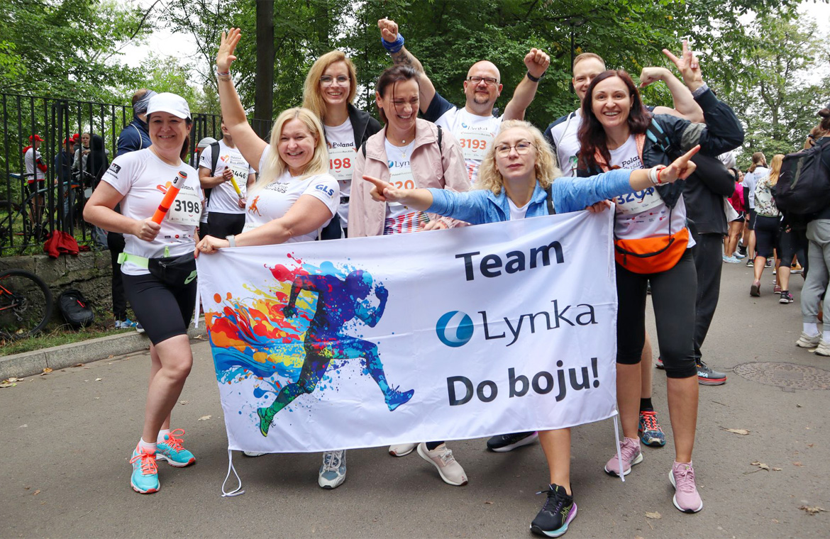 Uśmiechnięta drużyna Lynka w koszulkach startowych pozuje z banerem „Team Lynka – Do boju!" po biegu.